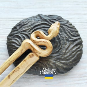 Könnte beinhalten: Ein Holzhaarstecker, der in Form einer Schlange geschnitzt ist. Der Stecker ist hellbraun mit dunkelbraunen Holzmaserungen. Die Schlange ist eingerollt, mit dem Kopf nach oben gerichtet. Der Stecker liegt auf einer dunkelbraunen Holzoberfläche.