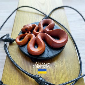 Może przedstawiać: Naszyjnik z drewnianym wisiorem przedstawiającym czerwono-pomarańczowego węża. Wisiorek jest okrągły i ma czarne tło. Naszyjnik wykonany jest z czarnego sznurka.