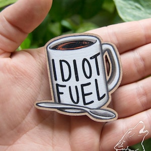 May include: A wooden coffee mug with a white background and black text that reads "Idiot Fuel". The mug is filled with brown coffee and has a white handle. A small wooden spoon is included.