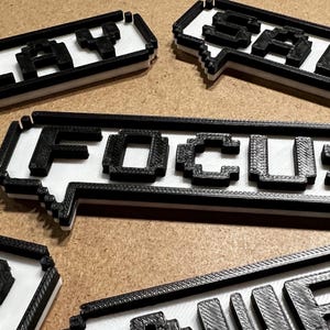May include: Several 3D printed rectangular speech bubble signs with black pixelated lettering on a white background. The signs feature the words SLAY, SALT, FOCUS, UP, and POWER, each rendered in a blocky, retro-inspired font style.
