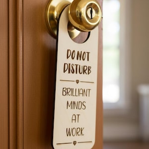 May include: A light-coloured wooden door hanger suspended from a polished brass doorknob. The hanger features the laser-engraved text DO NOT DISTURB, followed by a diamond icon, and the text BRILLIANT MINDS AT WORK, finished with another diamond icon.
