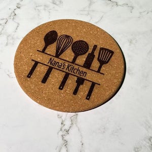 May include: A round cork coaster featuring a dark brown laser-engraved design of various kitchen utensils including a spoon, whisk, strainer, rolling pin, and spatula, with the text Nana's Kitchen centered horizontally across the utensils.