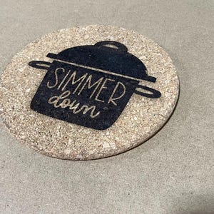 May include: A round, tan-colored cork coaster featuring a black graphic of a cooking pot with a lid. The text "SIMMER down" is printed in a light, cursive font inside the black pot graphic, creating a clear contrast against the textured cork surface.