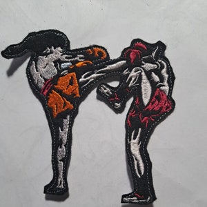 Op de afbeelding: Gestikt patch met twee figuren in een vechtpose. De figuur links draagt ​​oranje shorts en de figuur rechts draagt ​​rode shorts. Beide figuren bevinden zich in een dynamische pose met gestrekte benen en opgeheven armen.