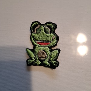 May include: Embroidered patch featuring a smiling green frog with a brown belly and black outline. The frog has large, friendly eyes and a red mouth. This patch is suitable for decorating clothing, bags, or other fabric items.