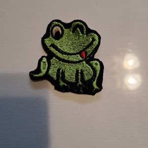 May include: Green embroidered frog patch with a black outline and a red tongue sticking out. The frog is sitting with its legs crossed and has a happy expression.