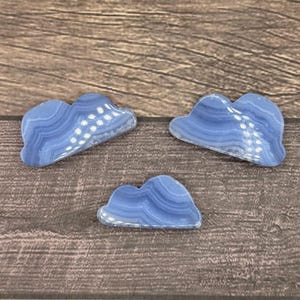May include: Three blue agate cloud-shaped stones on a wooden surface. The stones are smooth and have a natural, organic shape.