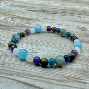 Puede incluir: Un brazalete de cuentas multicolores con cuentas azules, verdes, moradas y blancas. El brazalete est&aacute; hecho de piedras naturales y est&aacute; colocado sobre una superficie de madera.