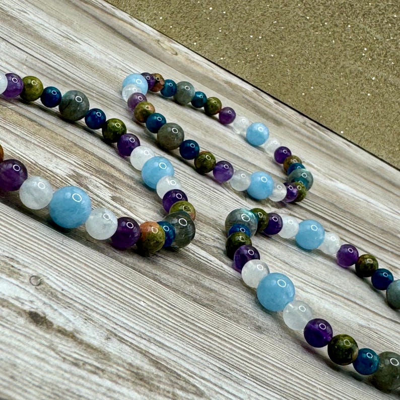 Puede incluir: Tres pulseras de cuentas con piedras de varios colores, incluyendo azul, morado, blanco y verde. Las pulseras est&aacute;n dispuestas en una fila sobre una superficie de madera.