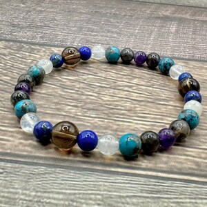 May include: A beaded bracelet featuring blue, brown, white, and purple stones. The bracelet is crafted from natural stones and is designed to be worn on the wrist.