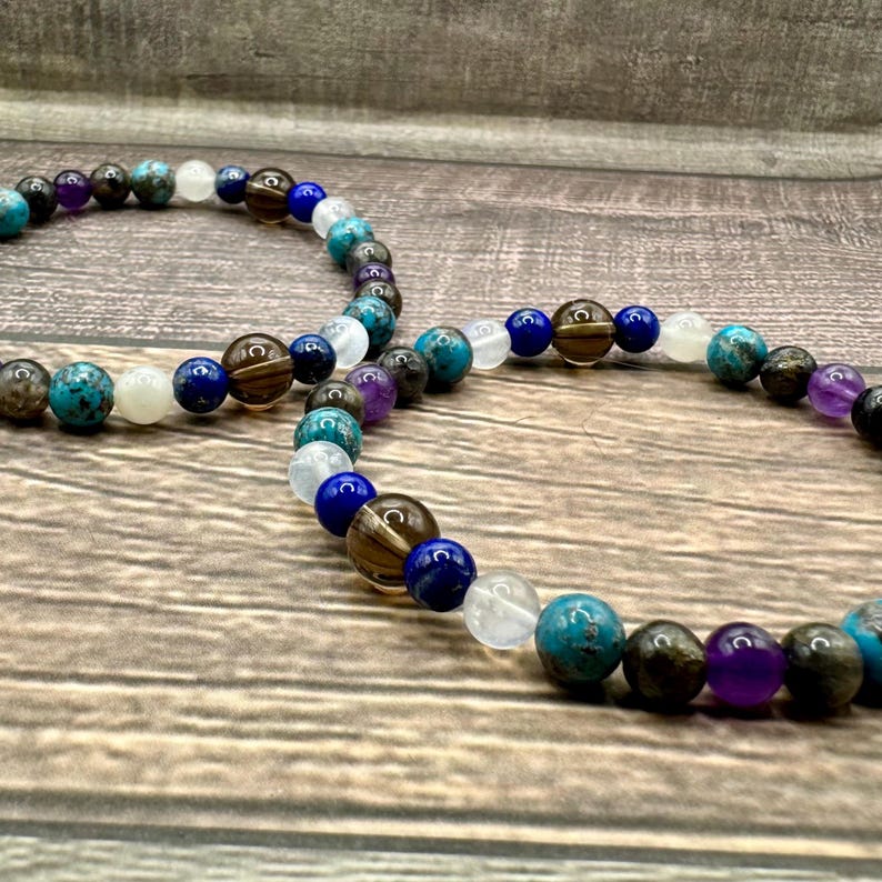 May include: Two beaded bracelets featuring brown, blue, white, and purple beads. The bracelets are crafted from natural stones and are layered on top of one another.