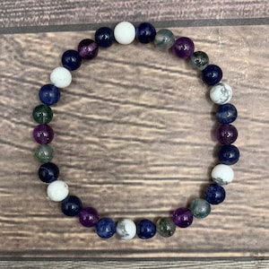 May include: A bracelet made with white, purple, and blue beads. The beads are round and have a smooth finish. The bracelet is on a wooden surface.