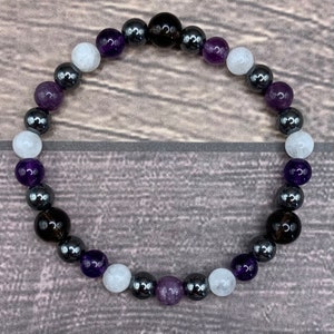 May include: A beaded bracelet with alternating amethyst, white, and black beads. The bracelet is made with natural stones and is designed to be worn on the wrist.