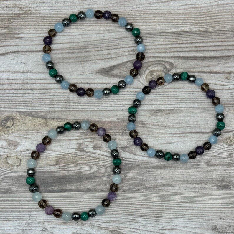 Pode incluir: Tr&ecirc;s pulseiras de contas com pedras de v&aacute;rias cores, incluindo azul, verde, marrom e roxo. As pulseiras est&atilde;o dispostas em c&iacute;rculo sobre uma superf&iacute;cie de madeira.