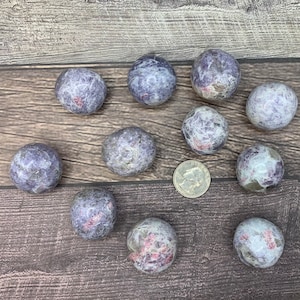 May include: A collection of ten tumbled Lepidolite spheres, ranging in size from small to medium. The spheres are a mix of purple, grey, and white with hints of pink. A US quarter is included for scale.