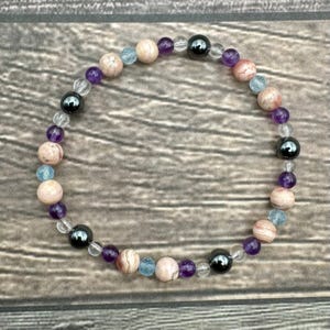 May include: A bracelet made of various gemstones, including amethyst, aquamarine, and hematite. The bracelet is strung on clear elastic.