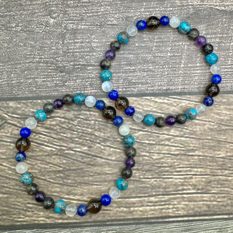 May include: Two beaded bracelets featuring a combination of blue, brown, grey and white beads. The bracelets are crafted from natural stones and are layered on top of one another.