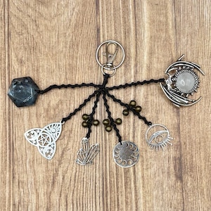 May include: A keychain featuring a silver-tone metal ring and clasp attached to multiple black knotted cords. The cords hold a dark faceted stone, a pentacle charm, a crystal cluster charm, small bronze beads, a moon phase disc, an eye charm, and a dragon.