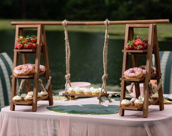Puede incluir: Dos soportes de madera de varios niveles con un columpio colgado entre ellos. Los soportes están decorados con cupcakes, donuts y fresas. Los soportes están colocados sobre un mantel rosa con hojas verdes.