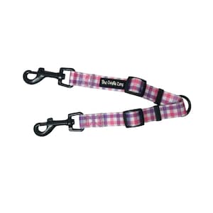Op de afbeelding: Een roze en paarse geruite hondenriem met twee zwarte metalen clips en een zwarte gesp. De riem heeft een zwart label met de tekst "The Crafty Dog".