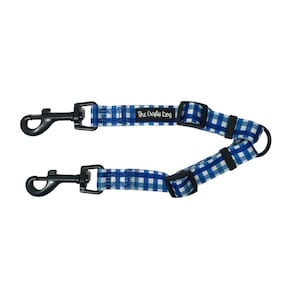 Op de afbeelding: Blauw-wit geblokt hondenriem met twee zwarte metalen clips en een zwarte gesp. De riem heeft de tekst "The Crafty Dog" erop gedrukt.