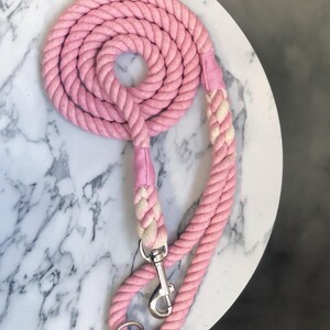 Può includere: Un guinzaglio rosa per cani realizzato con una corda spessa e ritorta. Il guinzaglio è arrotolato a spirale e presenta un moschettone e un anello in metallo argentato. L'impugnatura e la sezione vicino al moschettone sono di colore più chiaro.