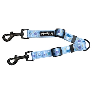 Op de afbeelding: Een lichtblauwe hondenriem met een herhalend patroon van paarse en blauwe vormen. De riem heeft twee zwarte metalen clips en een zwarte metalen ring. De tekst "The Crafty Dog" is gedrukt op een zwart label.