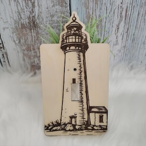 May include: A wooden light switch cover featuring a detailed engraving of a lighthouse. The lighthouse is depicted with intricate lines and shading, set against a light wood background. The design includes the lighthouse tower, a small adjacent building, and a rocky base.