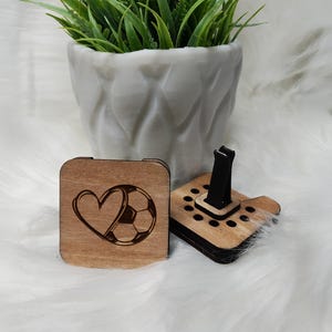 May include: A wooden car air freshener with a black clip. The front of the air freshener has a design of a heart and a soccer ball.
