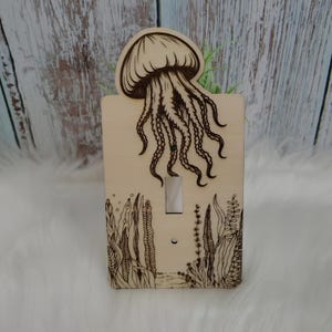 May include: A wooden light switch cover featuring a detailed engraved design of a jellyfish. The jellyfish is at the top, with its tentacles extending down. The bottom of the cover has an engraved underwater scene with plants. The switch is silver.