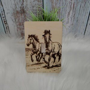 May include: Wooden light switch cover featuring a detailed engraving of two horses running. The horses are depicted in a dynamic pose, with flowing manes and tails. The cover has a natural wood grain finish and a standard light switch opening.