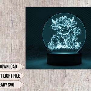 May include: A white night light with a cute cartoon highland cow design. The night light is on a black base and has a clear acrylic panel. The cow is surrounded by flowers.