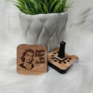 May include: A wooden car air freshener with a retro woman illustration and the text "Another Brilliant Idea, I'm Sure". The air freshener has a black clip to attach to a car vent.
