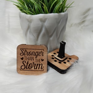 以下が含まれることがあります： 木製の車用携帯電話ホルダーとコースター。 ホルダーは黒い電話グリップと木製のベースを備えています。 正方形のコースターには「Stronger Than The Storm」の文字とハートのシンボルが刻まれています。