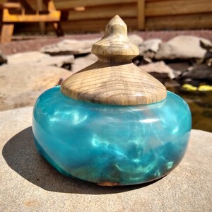 May include: A teal blue resin trinket box with a wooden lid. The lid has a rounded top and a wood grain pattern. The box is sitting on a rock.