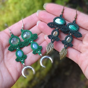 May include: Two pairs of handmade earrings. One pair features green macrame with moon charms and light blue and clear stones. The other pair has green macrame, leaf charms, and green and clear stones. The background is a blurred green.