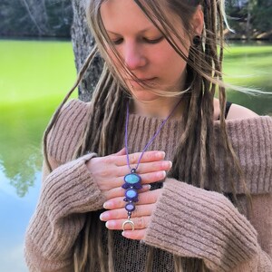 Op de afbeelding: Een handgemaakte ketting met een paars koord heeft een hanger met turquoise, lichtblauwe en paarse stenen. De hanger hangt aan een paars koord en heeft een zilveren halve maan bedel aan de onderkant. De ketting wordt gepresenteerd tegen een natuurlijke buitenachtergrond.