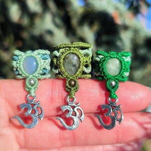 Op de afbeelding: Drie macraméringen in groen- en blauwtinten, elk met een edelsteen en een zilveren Om-bedel. De ringen zijn handgemaakt met ingewikkelde knopen, die een bohemien stijl laten zien. De edelstenen variëren in kleur en vorm.
