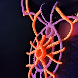 May include: A close-up of a neon orange and white pendant necklace. The necklace has a circular design with radiating lines and a central circular element. The background is dark, with hints of purple and blue.