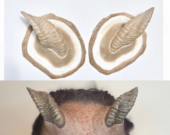 May include: Two beige, textured, molded horns with a white interior. The horns are shown on a white surface. A person is wearing the horns on their forehead.
