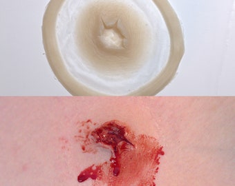 May include: A close-up of a fake wound, a round, flesh-colored silicone mold with a hole in the center, and a realistic-looking wound with red and brown paint applied to the mold.
