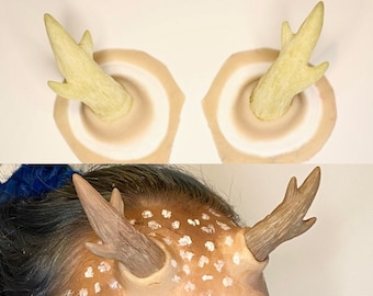 May include: Two beige deer antler headbands with a white inner circle. The antlers are made of a soft, flexible material and are designed to be worn on the forehead. The headbands are shown on a person's forehead, with white paint spots on their skin.