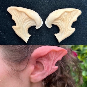 May include: A pair of beige, flexible, silicone prosthetic elf ears. The ears are designed to be applied to the side of the head and have a realistic, detailed texture.