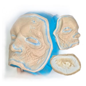 Peut inclure: Trois moules en silicone pour créer un masque facial détaillé et réaliste. Les moules sont beiges et ont une surface texturée. Le plus grand moule est partiellement rempli d'un liquide bleu.