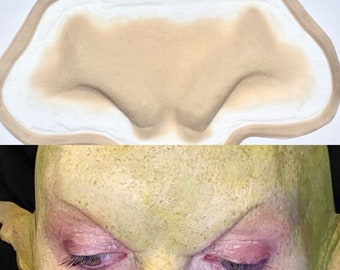 May include: A close-up of a prosthetic forehead appliance for special effects makeup. The appliance is made of a flexible, skin-like material and is designed to create a realistic, alien-like appearance. The appliance is shown in two states: unpainted and painted green.