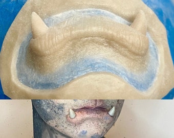 以下が含まれることがあります： 義歯の牙を作るための型をクローズアップした写真。型は薄いベージュ色のシリコン素材でできており、2つの尖った歯の形をしています。型は青い背景の上にあります。