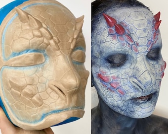 May include: A close-up of two different styles of makeup. One is a beige-colored prosthetic makeup piece with a textured, scaly design. The other is a blue and red makeup design with a similar scaly texture. Both designs are meant to resemble a reptilian creature.