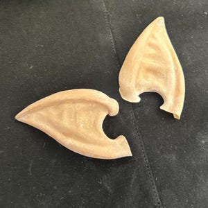 Könnte beinhalten: Zwei beige Elfenohren mit strukturierter Oberfläche. Die Ohren bestehen aus flexiblem Material und sind zum Tragen bestimmt. Die Ohren sind hellbraun und haben eine gebogene Form.