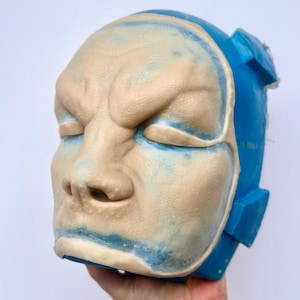 Peut inclure: Gros plan sur un moule en silicone beige pour le visage, entouré d'un moule en silicone bleu. Le moule pour le visage présente des détails précis, notamment les yeux, le nez et la bouche.