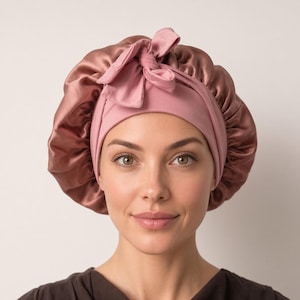 May include: A dusty rose satin sleep bonnet featuring a wide, matte-finish fabric band around the forehead and a decorative knotted bow detail at the top. The bonnet has a gathered, voluminous design with a smooth, shiny texture and soft sheen.